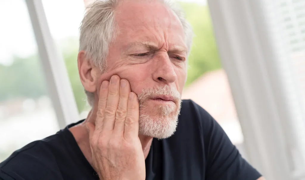 Older man with beard holding his hand to his jaw, appearing to be in pain or discomfort, possibly from a dental or tooth-related issue.