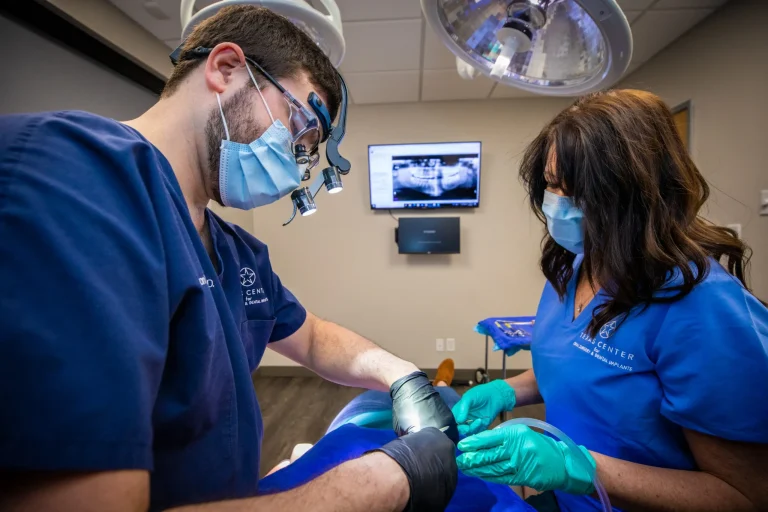 Oral surgeon performing dental implant surgery in a sterile operating setting, wearing masks, gloves, and surgical loupes while working with precision instruments.