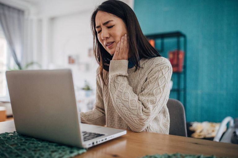 Woman experiencing TMJ jaw pain while holding the side of her face as she works on a laptop at home.