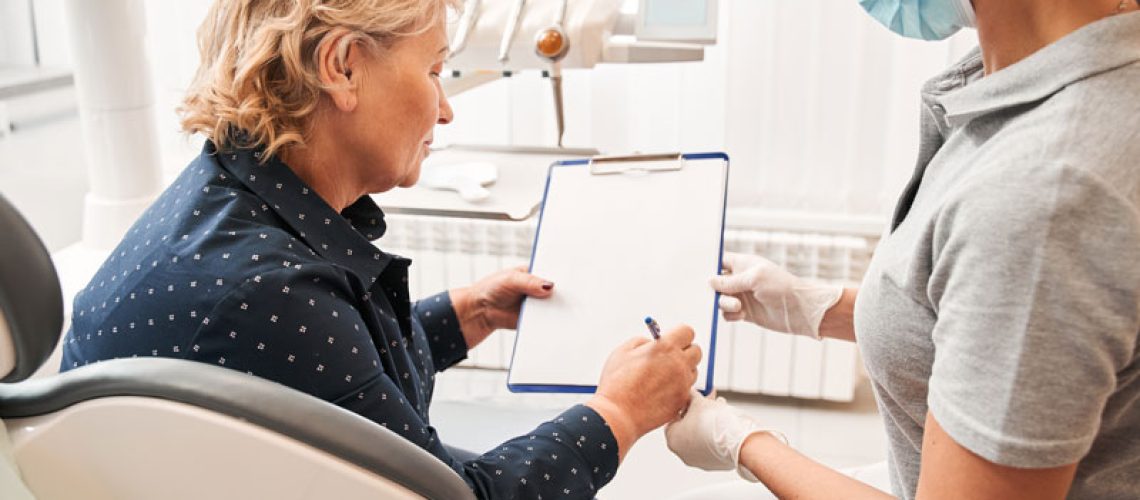 2024-05-Side-view-of-the-female-patient-with-a-pen-in-her-hand-signing-medical-application-form-of-her-new-dentist-sitting-in-front-of-her