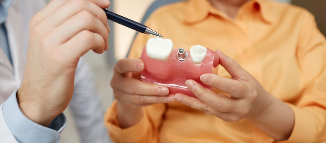 Closeup-of-unrecognizable-dentist-pointing-at-tooth-model-while-explaining-dental-implant-surgery-to-patient-in-clinic