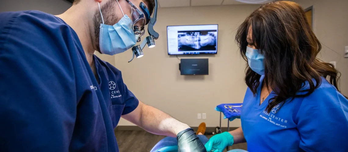 Oral surgeon performing dental implant surgery in a sterile operating setting, wearing masks, gloves, and surgical loupes while working with precision instruments.